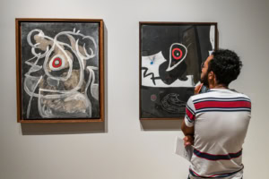 Un homme en polo rayé de noir, blanc et rouge observe deux peintures abstraites dominées par le noir, le blanc et le rouge.