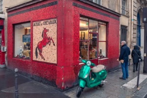 Boutique d’angle à façade rouge avec mosaïque représentant un cheval cabré et Vespa verte stationnée devant, à Paris.