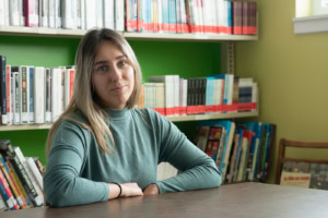 Une jeune femme aux cheveux blonds et aux yeux clairs est assise à une table dans la bibliothèque d'une école. Elle porte un chandail vert qui s'harmonise avec le mur du fond, créant une douce unité visuelle. Son regard, posé légèrement de côté, exprime une présence sereine et attentive. Derrière elle, des étagères remplies de livres colorés renforcent l'ambiance studieuse et intellectuelle du lieu.