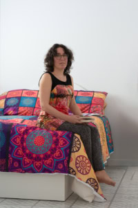 Jeune femme assise sur un lit dans une chambre, tenant un livre, avec couvre-lit et oreillers multicolores.