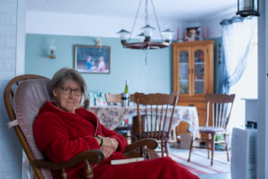 Femme âgée en robe de chambre rouge assise dans un fauteuil à bascule, dans un salon ouvert sur une salle à manger avec table, chaises en bois et vaisselier.