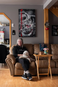 Portrait d’une femme assise dans un intérieur avec un petit chien sur les genoux, sous un tableau accroché au mur dans une pièce sobre.