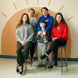 Photographie scolaire d'enfants ukrainiens et de leurs mères immigrés au Canada, à Tête-à-la-Baleine. Le groupe pose devant un décor mural représentant un soleil stylisé. Deux adolescentes sont assises sur des tabourets, vêtues de pulls contrastants, l'une en gris et l'autre en rouge. Un jeune garçon se tient debout, entouré par deux femmes, probablement ses proches, affichant une expression bienveillante. L'image évoque la solidarité et l'intégration dans un nouveau pays.