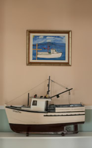Photographie d’une maquette de bateau posée sur une étagère sous une peinture encadrée représentant une embarcation, dans un intérieur à Tête-à-la-Baleine, Québec, Canada.