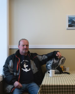 Portrait d’un homme assis à une table dans une auberge à Tête-à-la-Baleine, tenant un casque de motoneige, avec une tasse et un décor intérieur sobre.