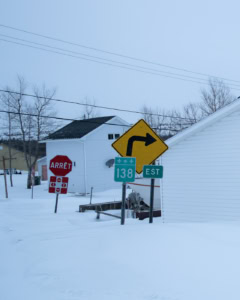 Panneaux de signalisation indiquant un virage à droite, la route 138 Est et un arrêt, devant des maisons blanches dans un paysage enneigé.