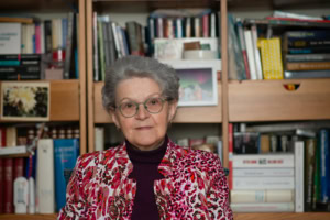 Femme âgée aux cheveux gris et aux lunettes rondes, devant une bibliothèque remplie de livres, portant une veste colorée sur un col roulé foncé.