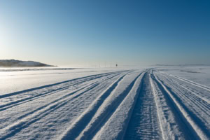 Traces de motoneige sur une étendue gelée dans la Baie Plate, à Tête-à-la-Baleine, sous un ciel bleu, avec des balises au loin et une colline enneigée à gauche.