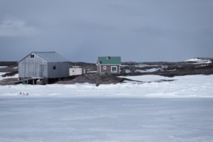 Quelques maisons dispersées sur l’île Anderson, au bord d’une étendue d’eau gelée, avec une cabane en bois sur pilotis et une maison au toit vert.