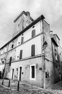 Bâtiment en angle à Arles mêlant ancienne maçonnerie d’église et façade enduite, avec volets fermés, rue pavée et tour en pierre en arrière-plan.