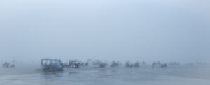 Plage panoramique dans un brouillard bleu dense. Des groupes de personnes restent installés sous des parasols ouverts, près de petites tentes ou sur des chaises de plage, dispersés entre les flaques de marée.