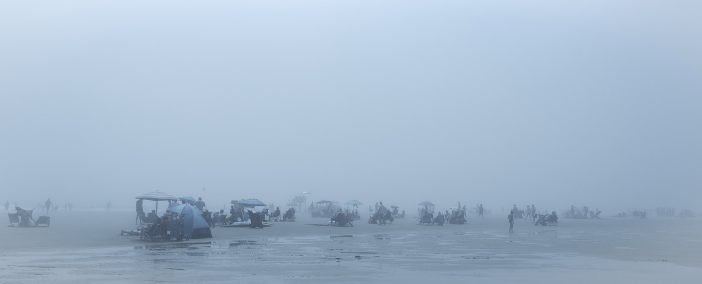 Plage panoramique dans un brouillard bleu dense. Des groupes de personnes restent installés sous des parasols ouverts, près de petites tentes ou sur des chaises de plage, dispersés entre les flaques de marée.