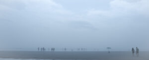 Plage panoramique dans un brouillard bleu dense. De nombreuses silhouettes se répartissent sur le rivage. À droite, un parasol se tient seul dans le sable, tandis qu’un couple marche dans sa direction au bord de l’image.