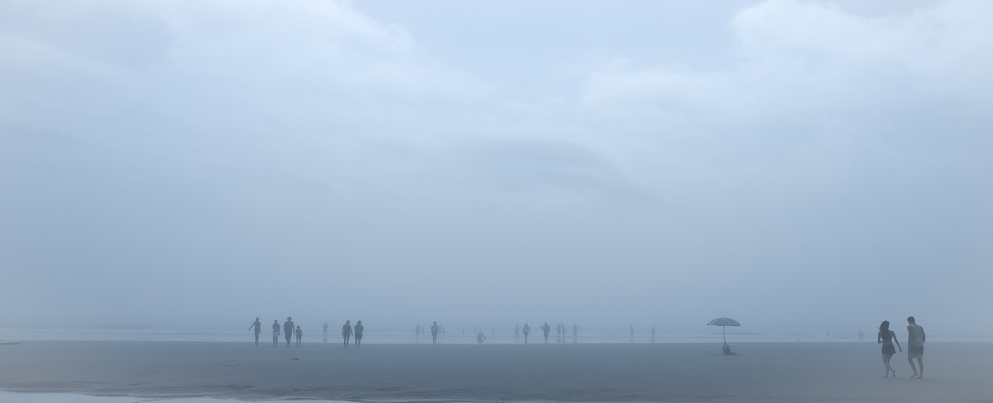 Plage panoramique dans un brouillard bleu dense. De nombreuses silhouettes se répartissent sur le rivage. À droite, un parasol se tient seul dans le sable, tandis qu’un couple marche dans sa direction au bord de l’image.