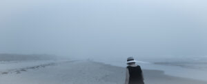 Plage panoramique dans un brouillard bleu dense. À droite, une femme vue de dos portant un chapeau blanc marche au bord de l’eau.