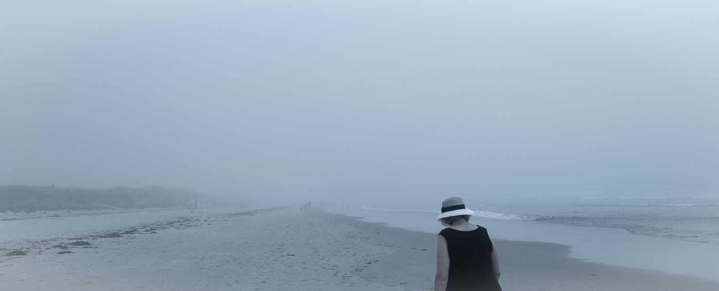Plage panoramique dans un brouillard bleu dense. À droite, une femme vue de dos portant un chapeau blanc marche au bord de l’eau.