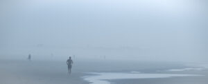 Plage panoramique dans un brouillard bleu dense. Au premier plan, un joggeur court vers nous, tandis que quelques silhouettes apparaissent plus loin sur le rivage.