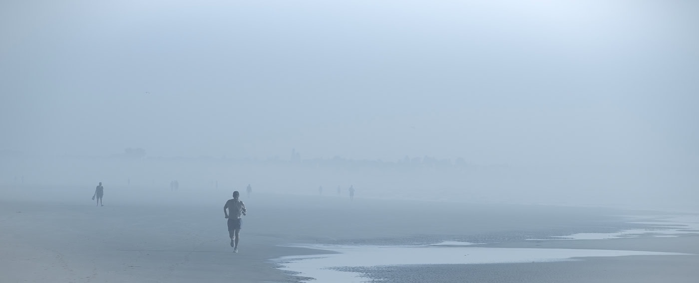 Plage panoramique dans un brouillard bleu dense. Au premier plan, un joggeur court vers nous, tandis que quelques silhouettes apparaissent plus loin sur le rivage.