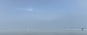 Plage panoramique dans un brouillard bleu. Une chaise haute de sauveteur se détache au centre-gauche, quelques silhouettes apparaissent au loin et un couple marche au bord de l’eau à droite.