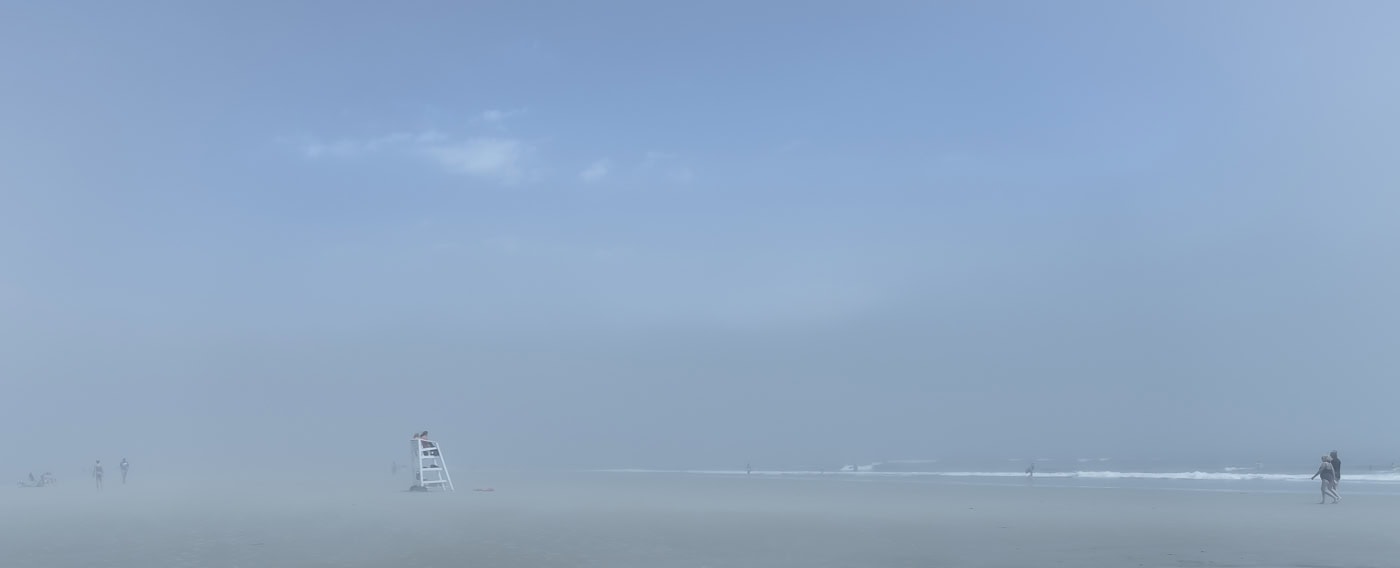 Plage panoramique dans un brouillard bleu. Une chaise haute de sauveteur se détache au centre-gauche, quelques silhouettes apparaissent au loin et un couple marche au bord de l’eau à droite.